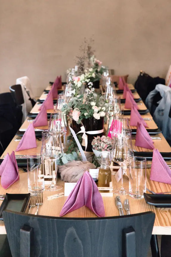 Langer Holztisch mit violett gefalteten Stoffservietten, Gläsern und natürlicher Blumendekoration, vorbereitet für eine Veranstaltung im Weingut Daniel & Monika Marugg.