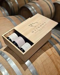 Halb geöffnete Holzweinkiste „Weingut Daniel & Monika Marugg" mit drei Pinot-Noir-Flaschen (Bovel, Intus) auf Weinfässern im Barrique-Keller