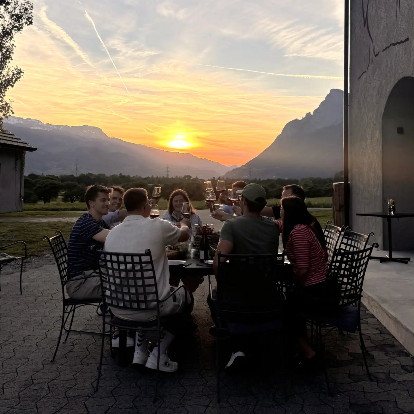 Eine Gruppe von Menschen sitzt bei Sonnenuntergang auf einer Terrasse im Freien an einem gedeckten Tisch, stößt mit Weingläsern an und genießt den Blick auf die Berge in der Abendstimmung.