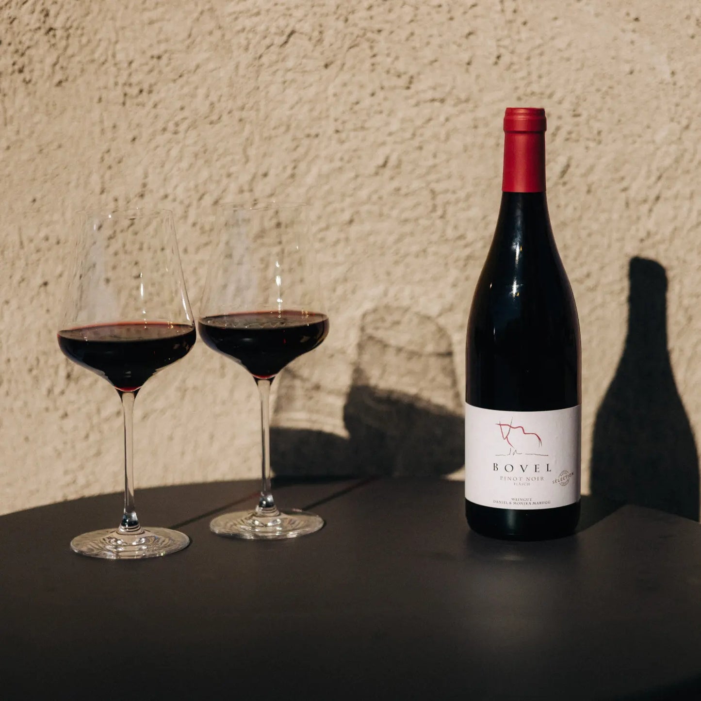 Zwei Gläser mit Rotwein und eine Flasche Pinot Noir mit Aufschrift Bovel und roter Kapsel stehen auf einem dunklen Tisch vor einer hellen, strukturierten Wand im Sonnenlicht.