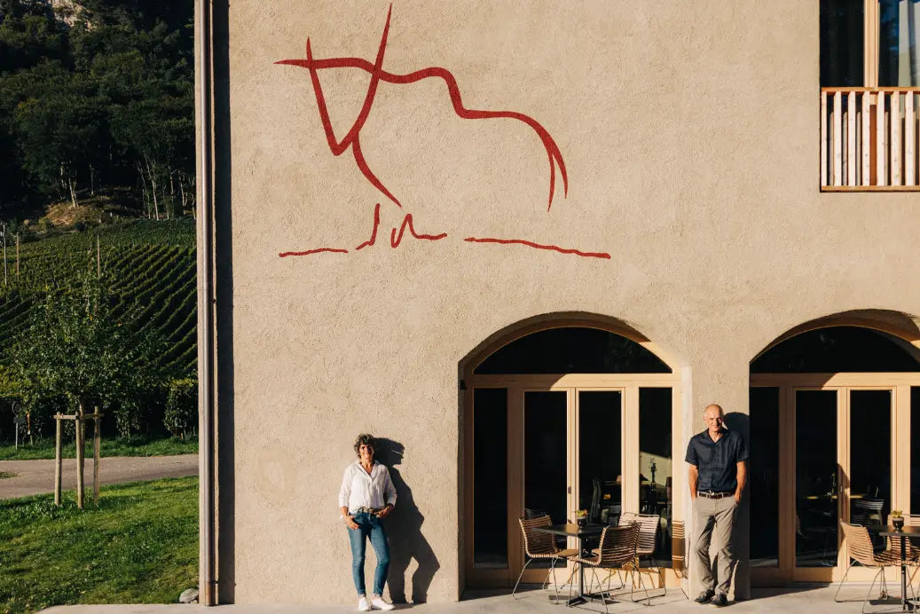 Zwei Personen vor dem Weingut-Gebäude mit charakteristischem roten Stier-Wandbild, Weinberge im Hintergrund