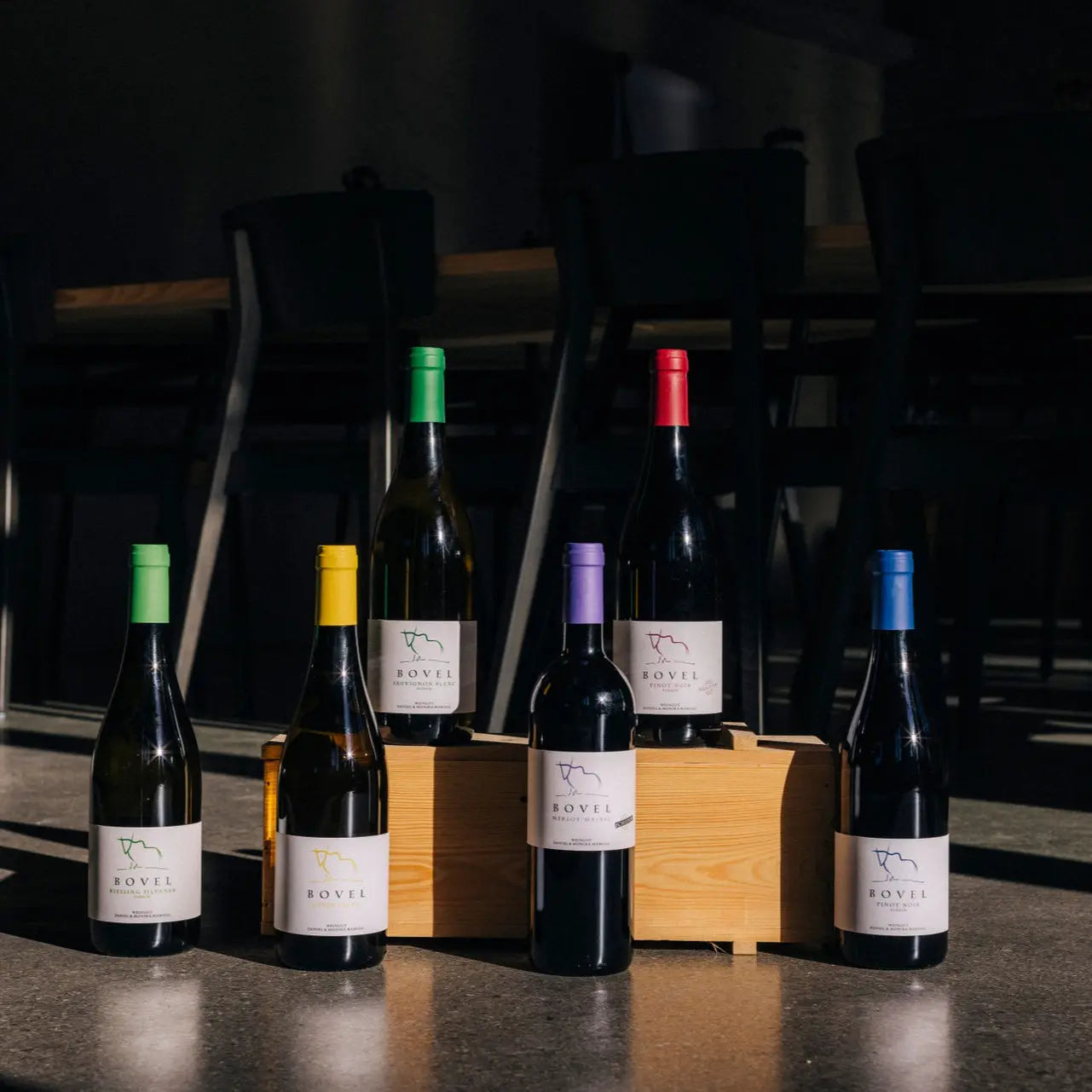 Sechs Flaschen der Bovel-Linie vom Weingut Marugg auf einem Tisch mit Holzkiste – Weiss- und Rotweine mit farbigen Kapseln, Fläsch, Bündner Herrschaft