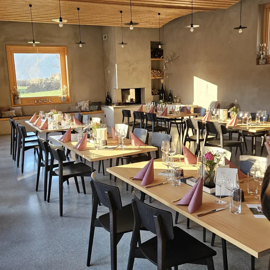 Heller, modern eingerichteter Gastraum mit mehreren gedeckten Holztischen und schwarzen Stühlen. Rosa gefaltete Servietten, Gläser und kleine Blumenarrangements stehen auf den Tischen. Im Hintergrund befinden sich ein Kamin, eine Sitzbank am Fenster und eine Holzdecke mit zahlreichen Pendelleuchten. Sonnenlicht fällt durch das große Fenster in den Raum.