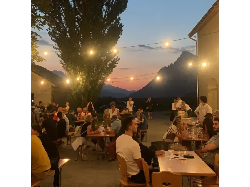 Abendstimmung im Innenhof des Weinguts Marugg in Fläsch: Gäste sitzen an langen Tischen unter Lichterketten, im Hintergrund die Silhouette der Bündner Berge im Sonnenuntergang.